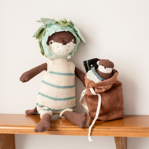 Clara Otter wearing striped swimsuit and pondweed swim cap sitting next to her dry bag backpack, which is open and has her mini otter peeking out